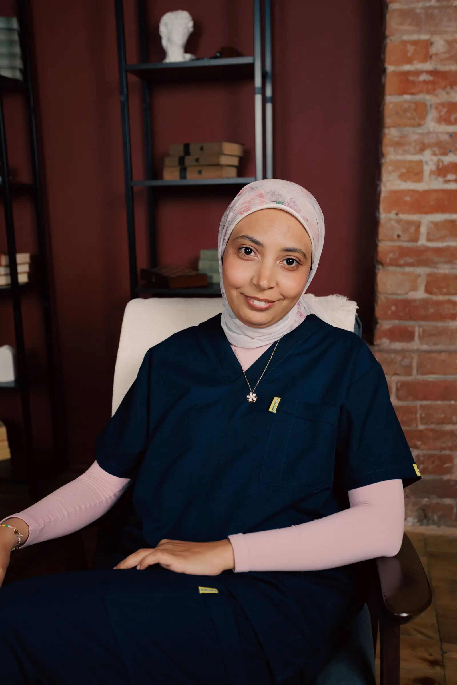 En vårdare i hijab och mörkblå arbetskläder sitter i en fåtölj och ler.